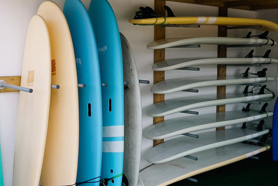 Surfboards of various colors and sizes stored under a wooden shelter.