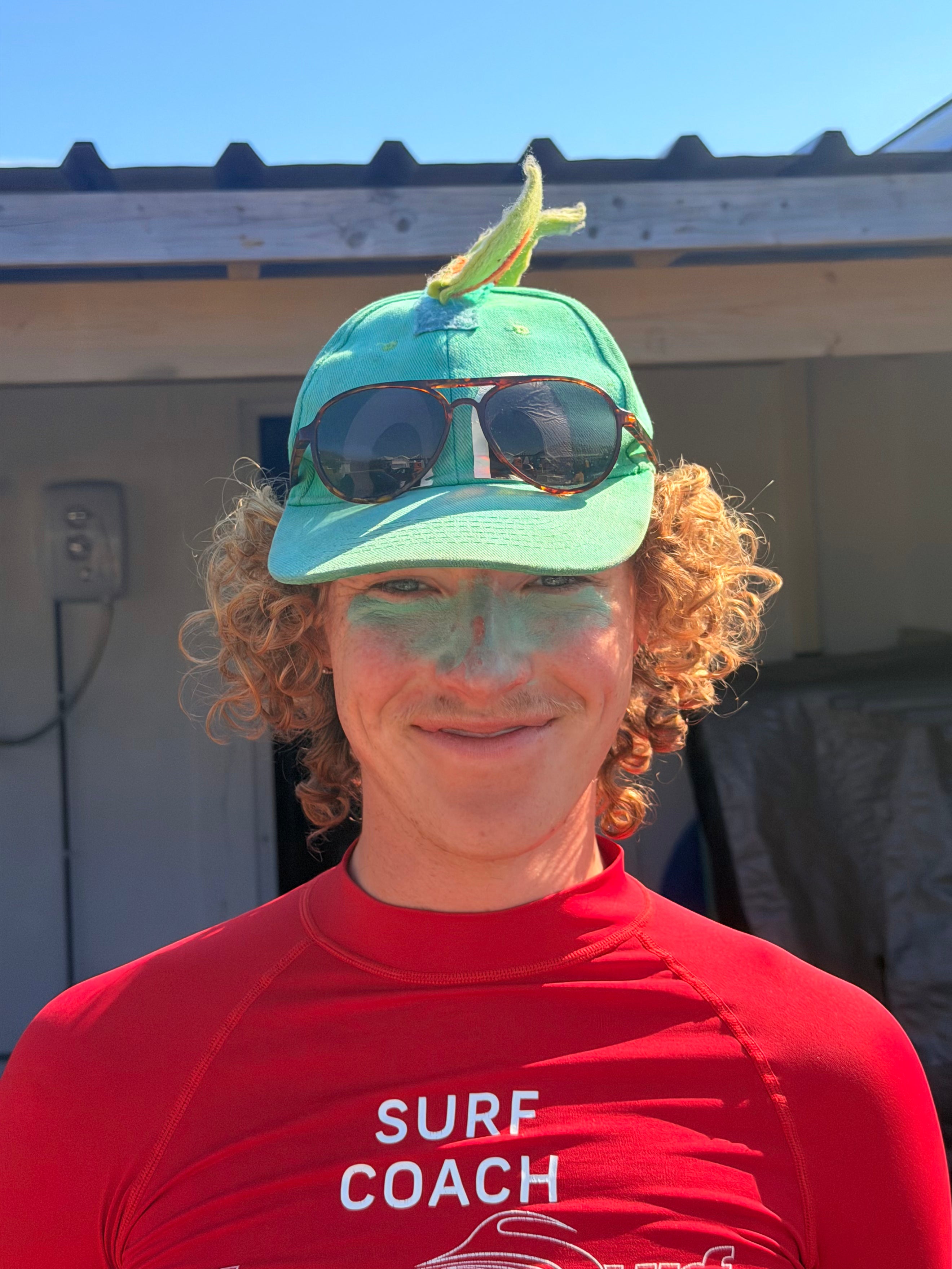Person wearing a red 'Surf Coach' shirt with a green cap and sunglasses on their head.